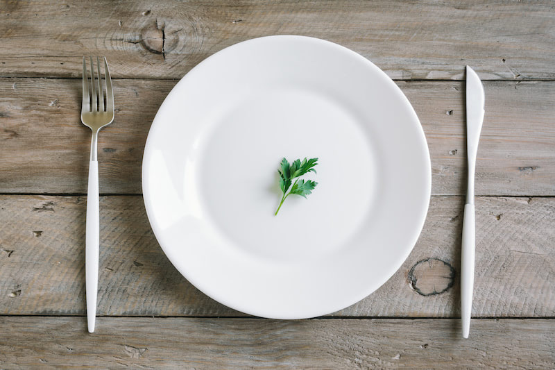 Table setting with white plate, modern cutlery (fork and knife) and little leaf of fresh parsley on wooden background, top view, copy space. Diet, fasting, vegan raw food concept.